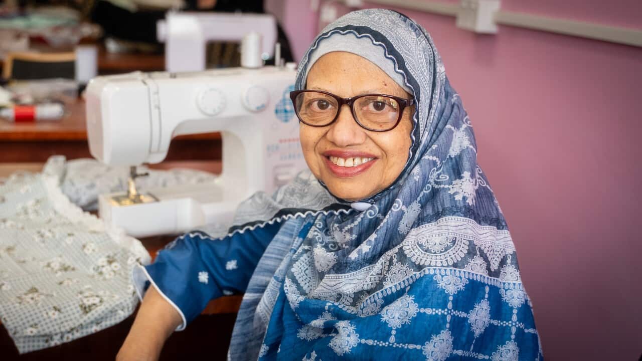 A woman in a blue shawl sits at a sewing machine smiling at the camera.