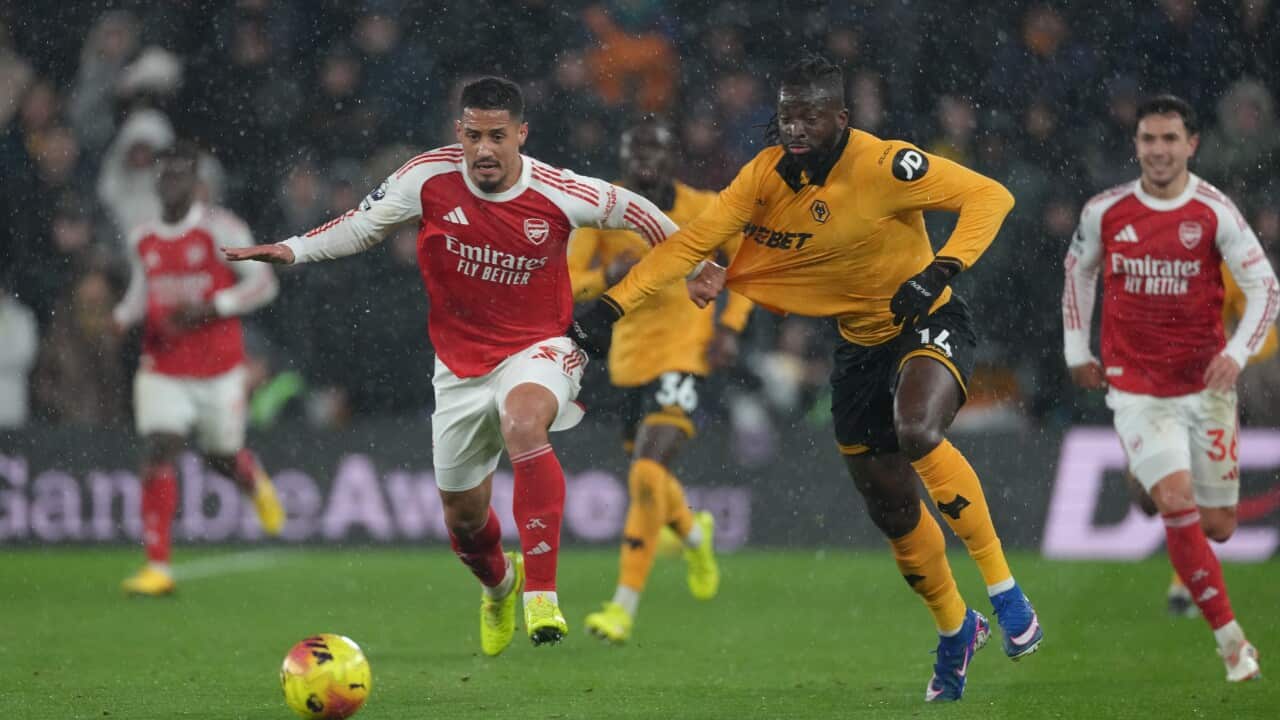 Two footballers chasing a ball on a field. The player on right is wearing an orange long-sleeved shirt and black shorts and the one on the left is wearing a red and long-sleeved white shirt and white shorts.