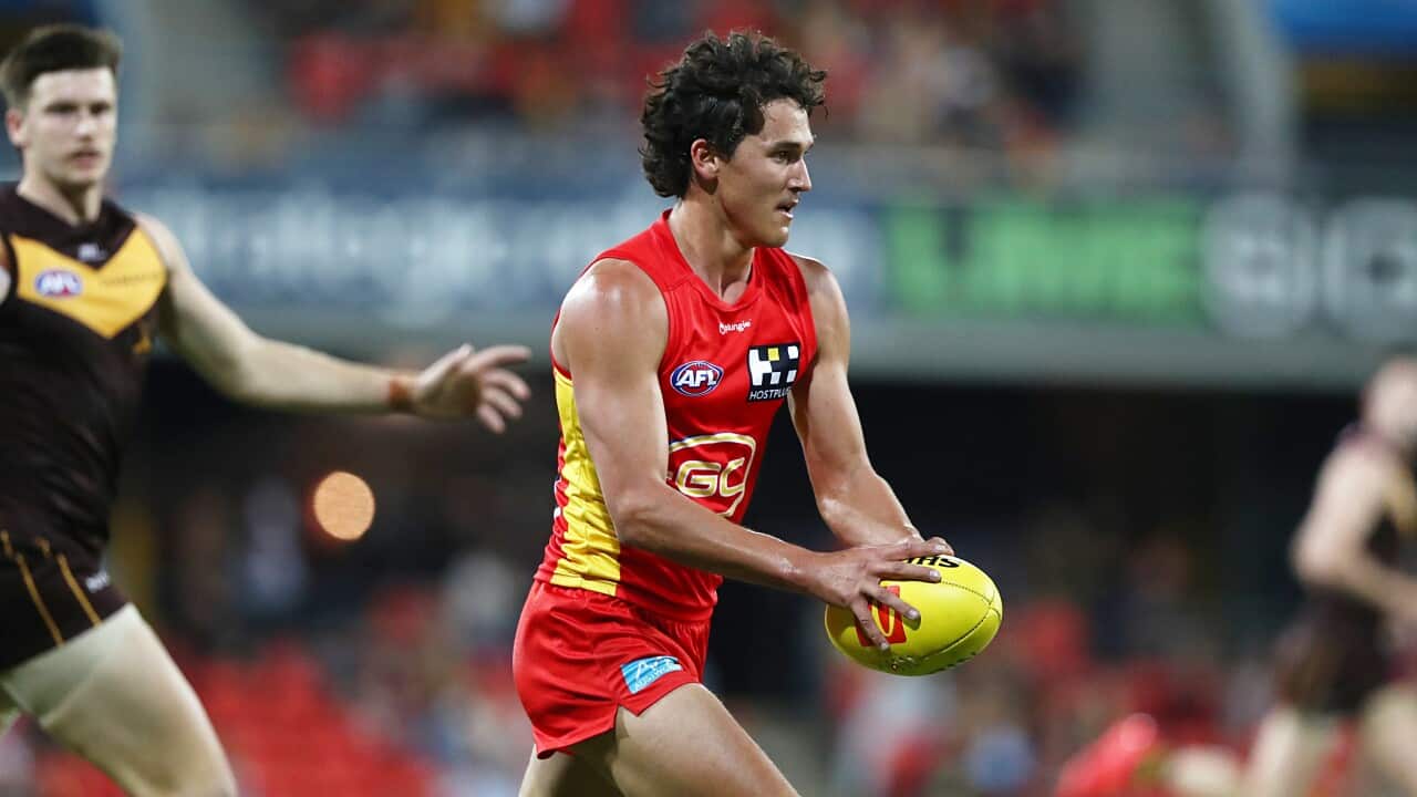 A young man with brown hair wearing a red AFL jersey runs with a yellow AFL ball in his hands. In the out-of-focus background, a crowd is visible.