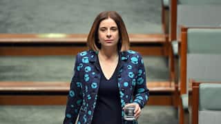 A woman in a blue patterned jacket holds a glass of water while walking down the steps of a parliamentary chamber.