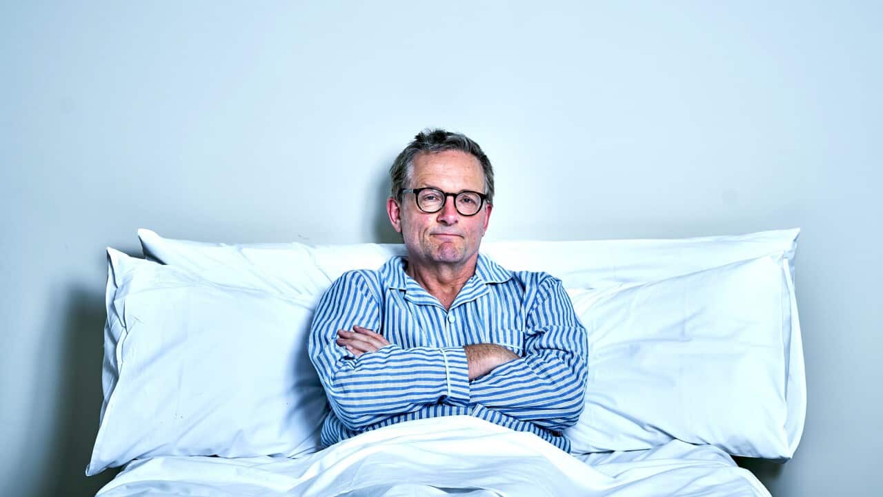 A man in striped pyjamas and glasses sits up in bed, leaning against the pillows. His arms are crossed and he has a wry, slightly glum smile.