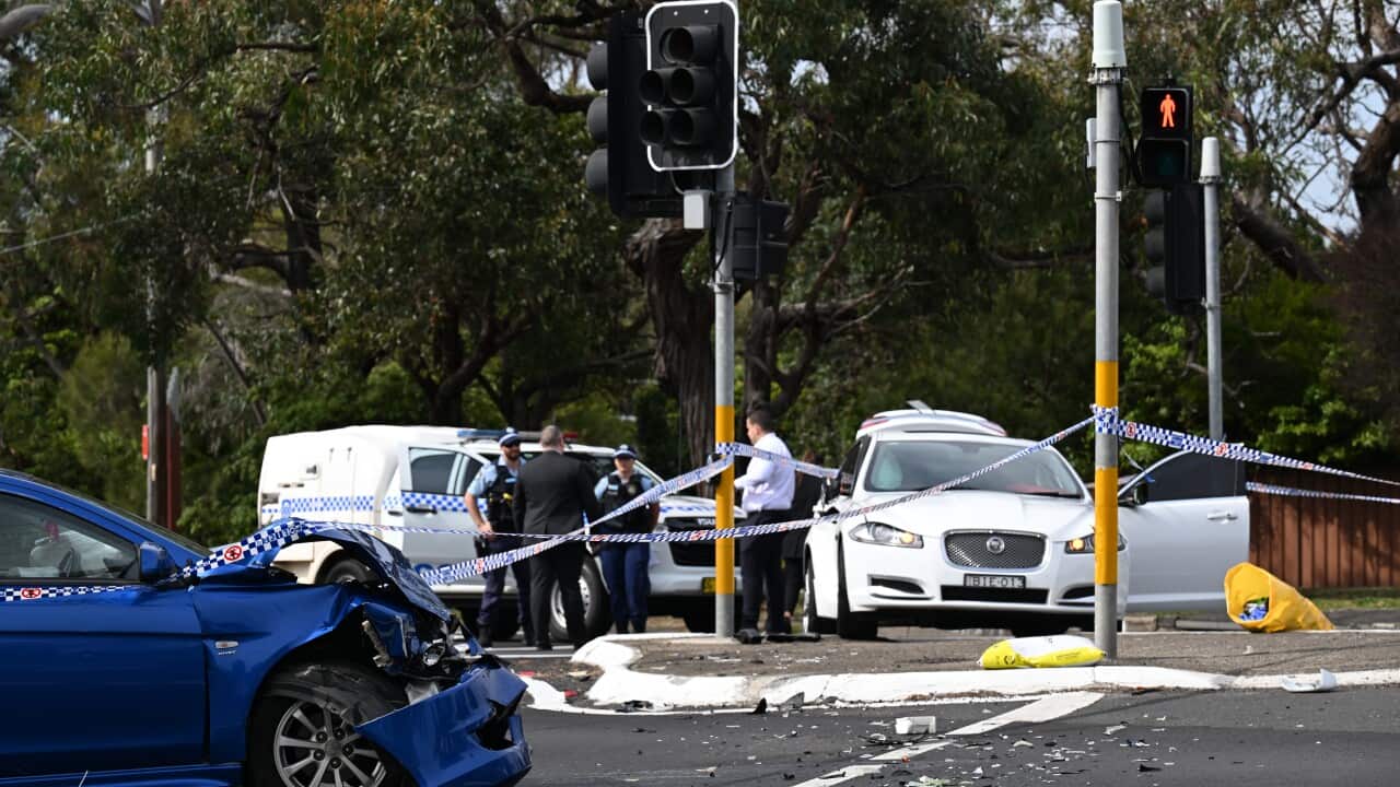 The scene following a car crash, with police tape and officers visible. Once car has a severely dented front bumper and there is shattered glass on the road.