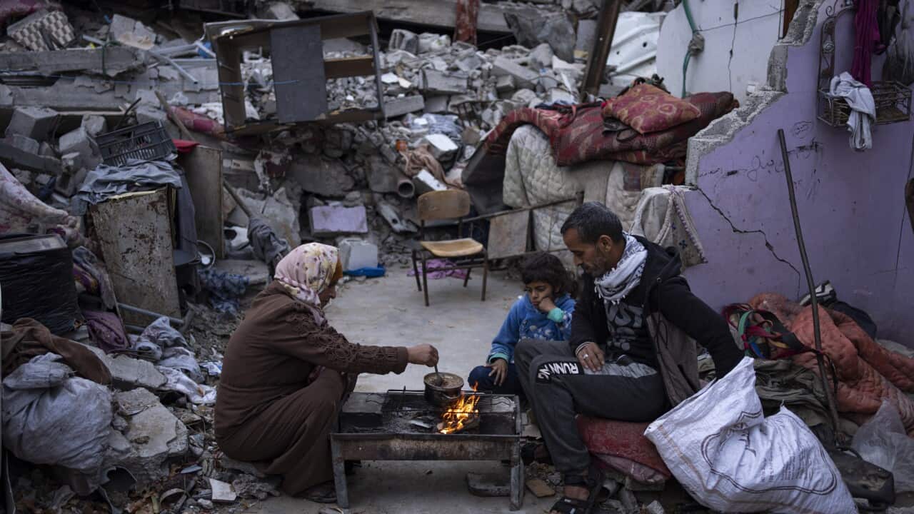 Two adults and a child sit next to a stove amid rubble.