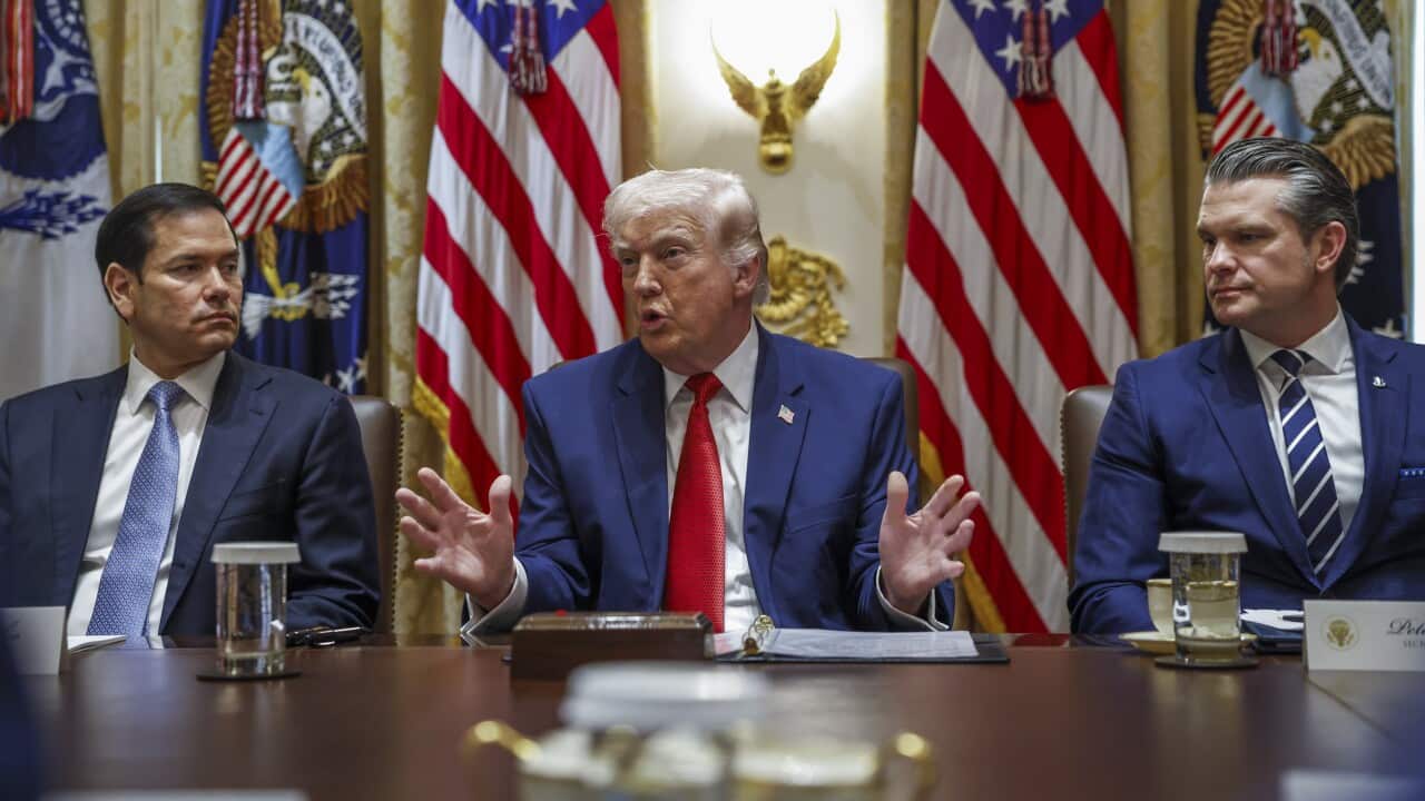 Donald Trump sits at a conference table flanked by Marco Rubio and Pete Hegseth, gesturing with his hands while speaking in front of several American flags.