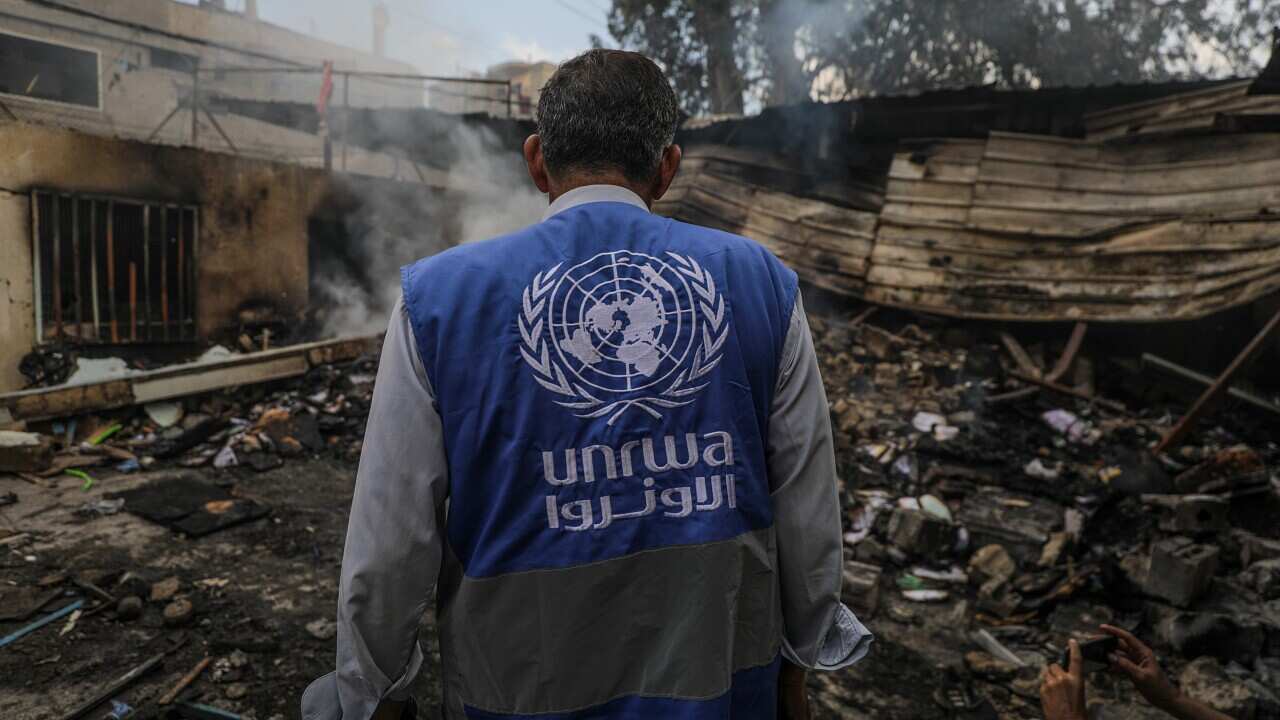 A man in a blue UNRWA jacket stands among the ruins with his back to the camera.