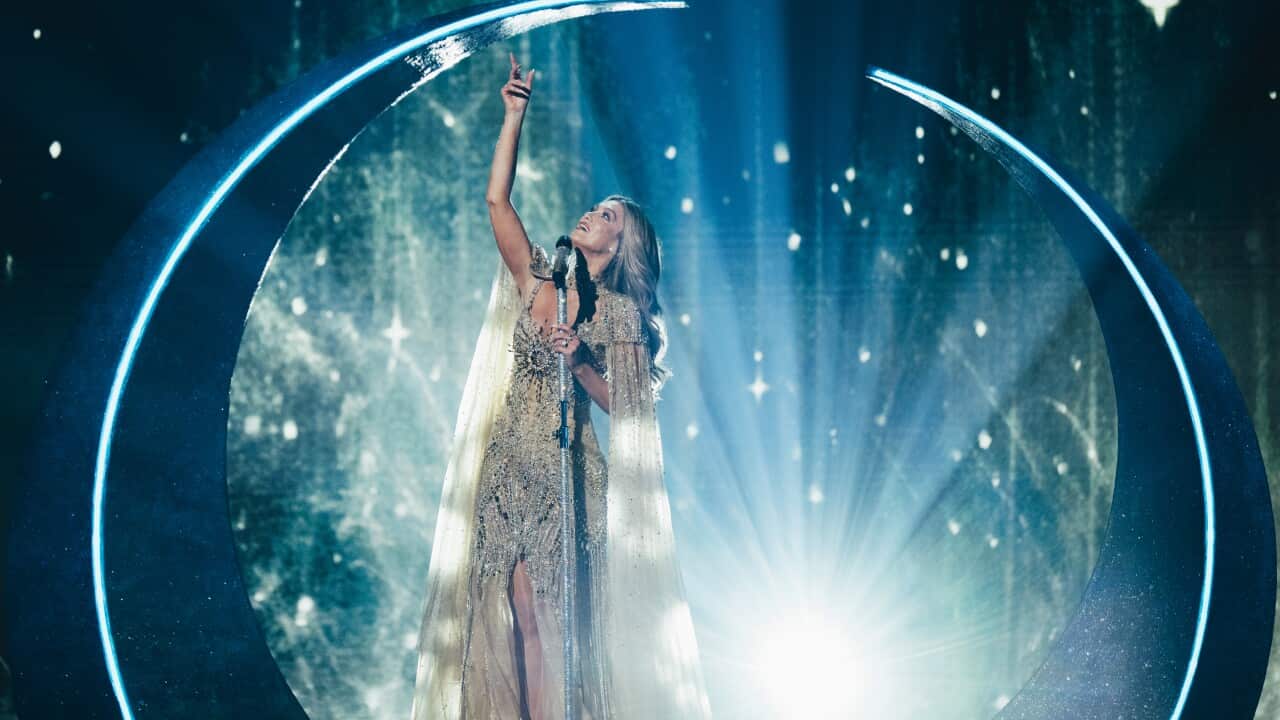 Delta Goodrem singing on stage. She is holding her right hand up and looking towards it as she holds a microphone stand with her left hand. The background is blue with a bright light and stars