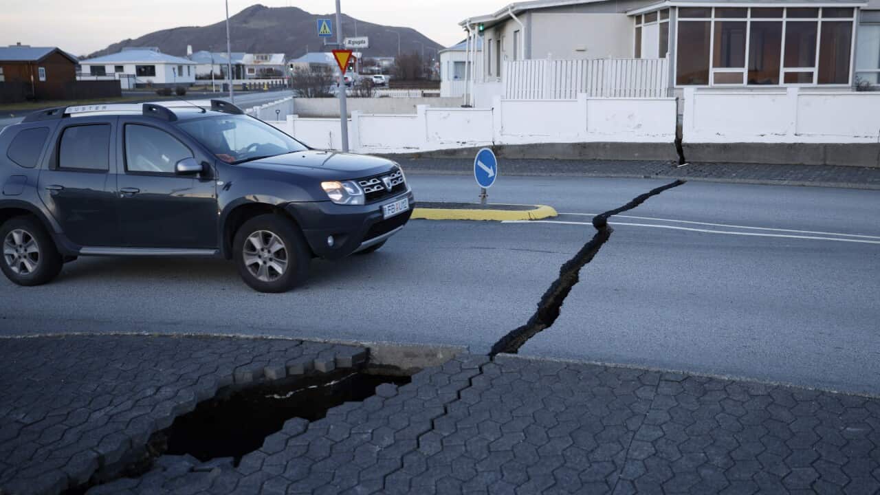 A small, black sports utility vehicle drives towards a large fissure in a road. The sidewalk and wall of a house is also cracked