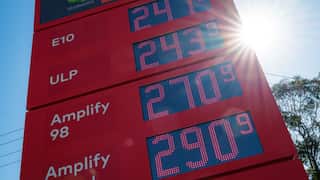 A red Woolworths petrol station price board displaying E10, ULP, Amplify 98 and Amplify Diesel prices, with bright sunlight flaring behind the sign.