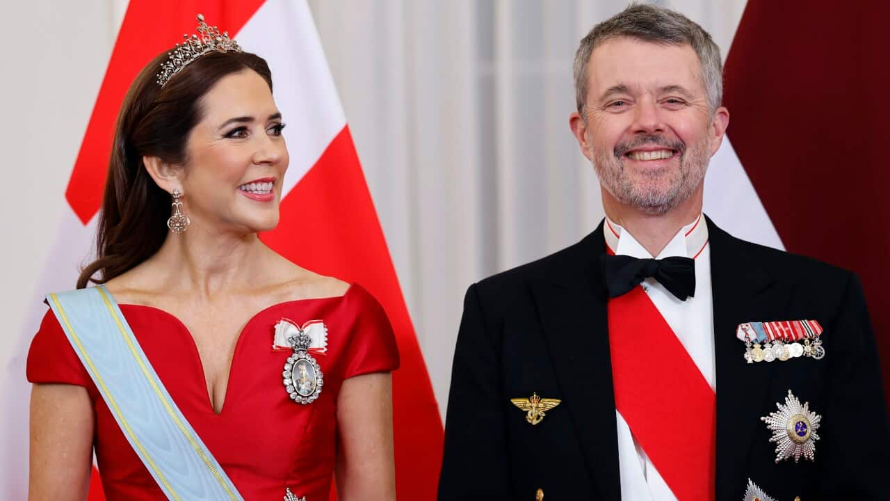 King Frederik X and Queen Mary of Denmark stand side-by-side in formal gala attire, including sashes, medals and a tiara, against a backdrop of the Danish flag.