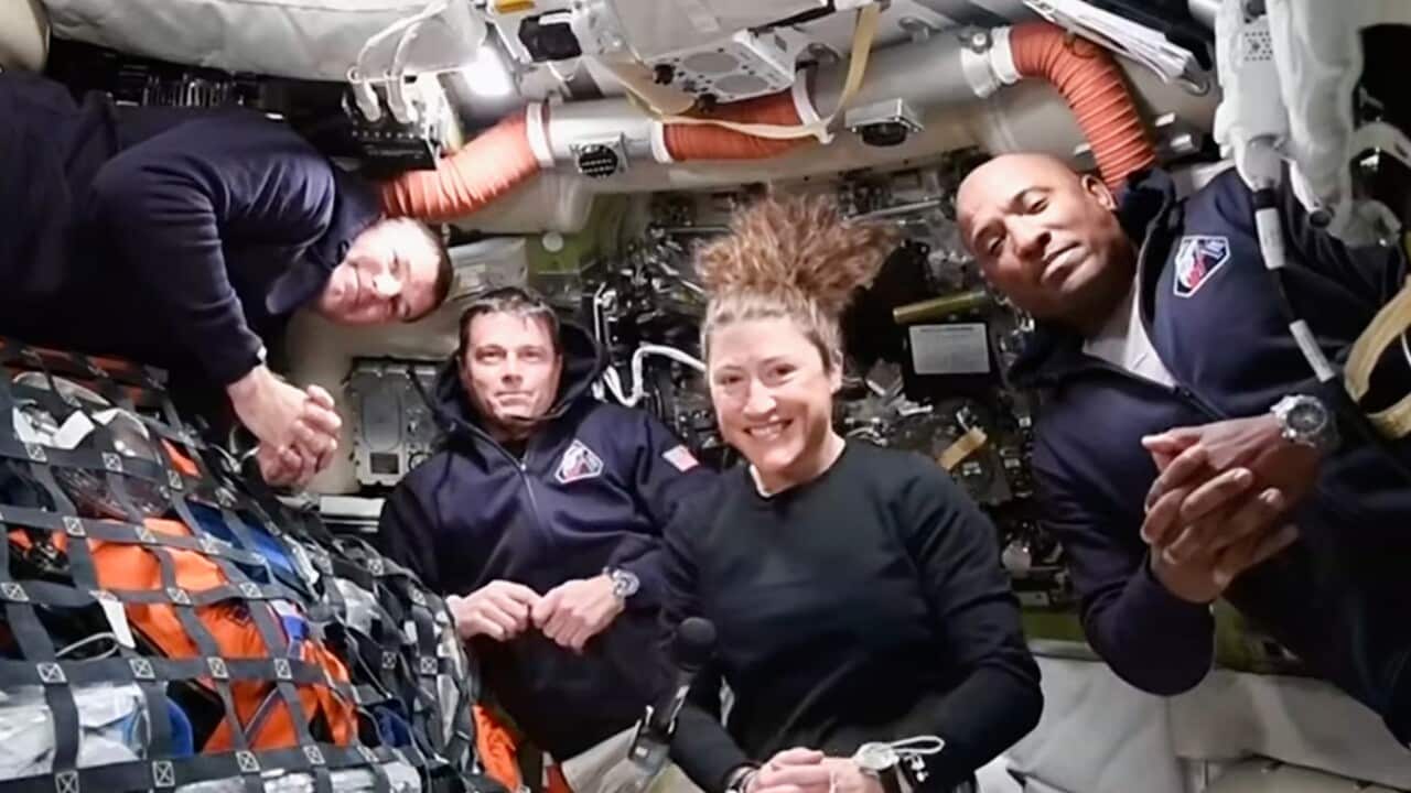 Four astronauts, including three men and one woman, pose together in the cramped, equipment-filled interior of a spacecraft while floating in microgravity.