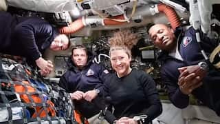 Four astronauts, including three men and one woman, pose together in the cramped, equipment-filled interior of a spacecraft while floating in microgravity.