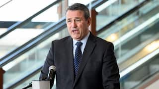 A man wearing a dark suit and blue tie speaks from a lectern bearing the Australian Government crest in front of a modern staircase.