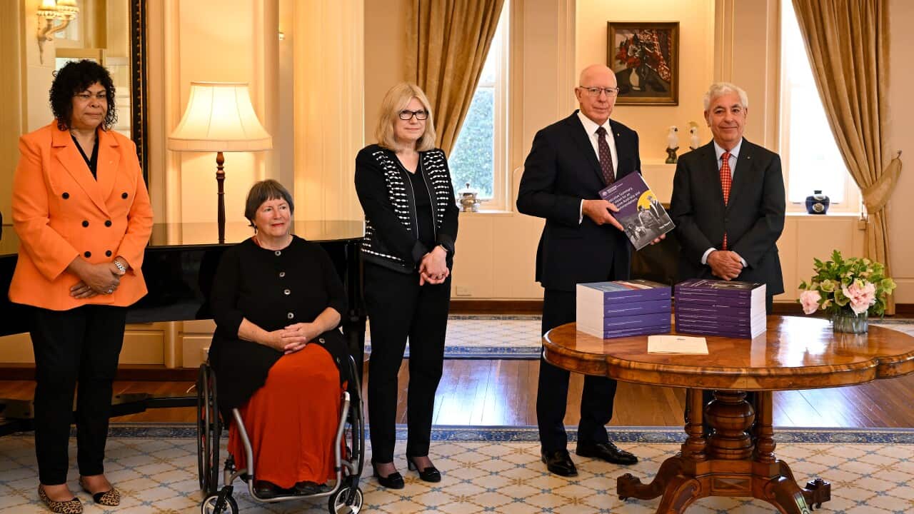 A group of formally dressed people stand in room with fancy decor. One is holding a copy of a report. There are stacks of the report on the table. One woman is in a wheelchair.