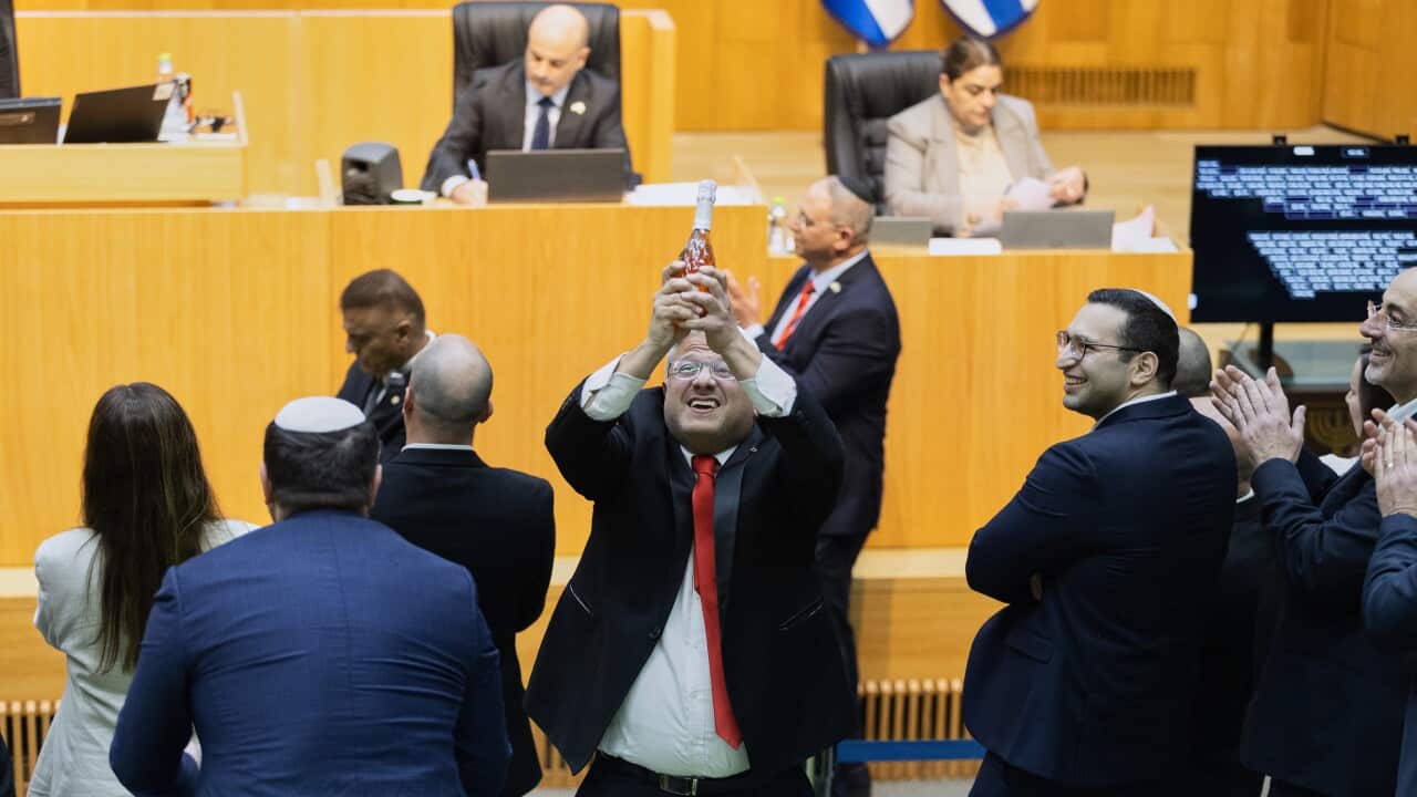 A man wearing a suit and a red tie holds his hands up as he celebrates inside a parliament chamber surrounded by other members