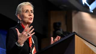 Micaela Cronin standing behind a lectern and speaking wearing a red scarf.