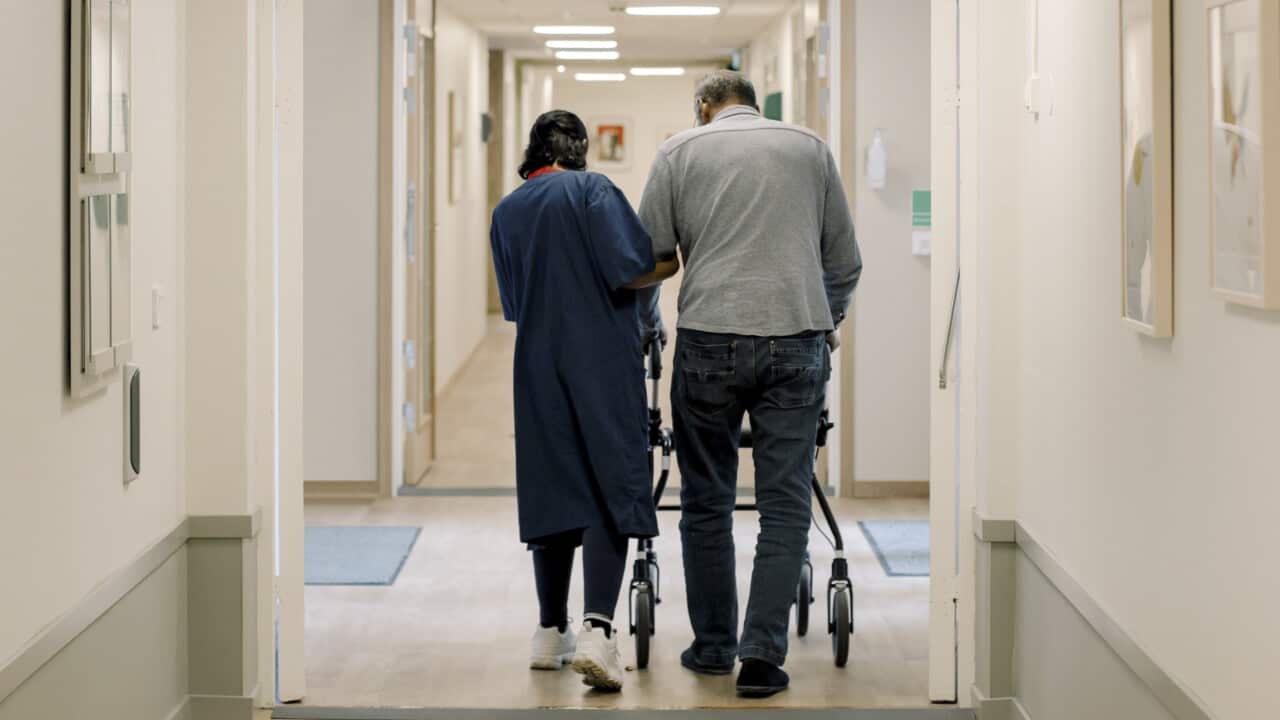 An aged care worker assisting a man with a walker down a corridor.