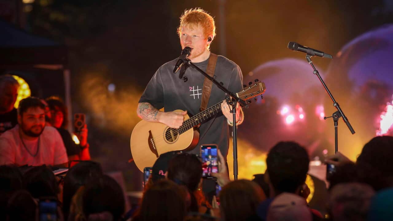 Musician Ed Sheeran sings into a microphone while playing an acoustic guitar on stage in front of a crowd during an outdoor concert.