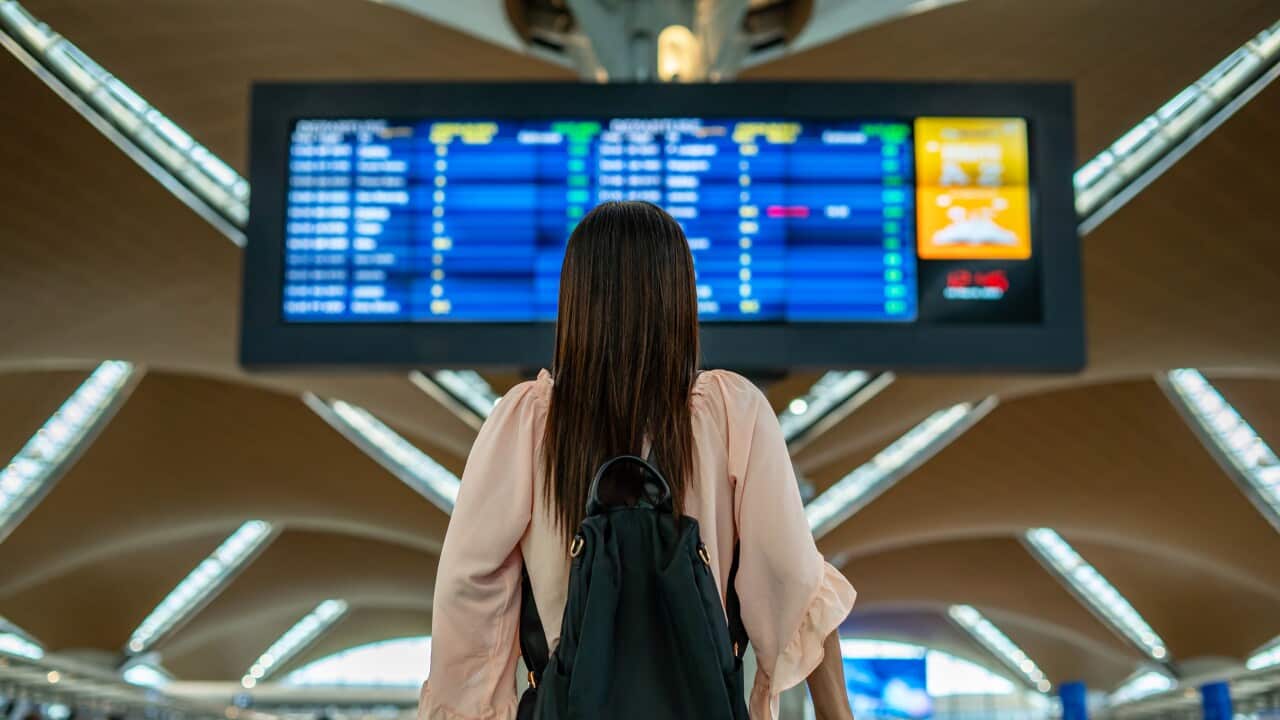 A woman standing in an airport wearing in a backpack holding a suitcase facing away from the camera