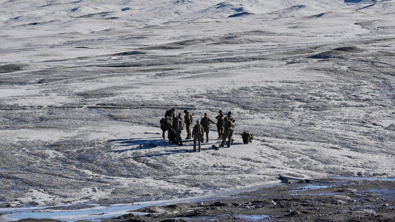 A group of military personnel stands in a huddle on a vast, rugged, and snow-dusted landscape under a clear sky.