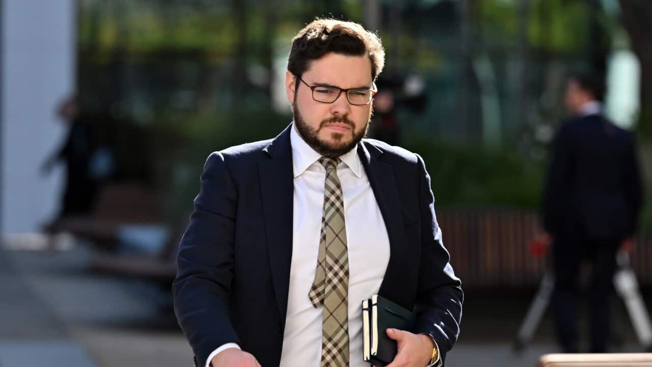 A man wearing a suit and carrying two diaries in his left hand, walks with his head bowed.