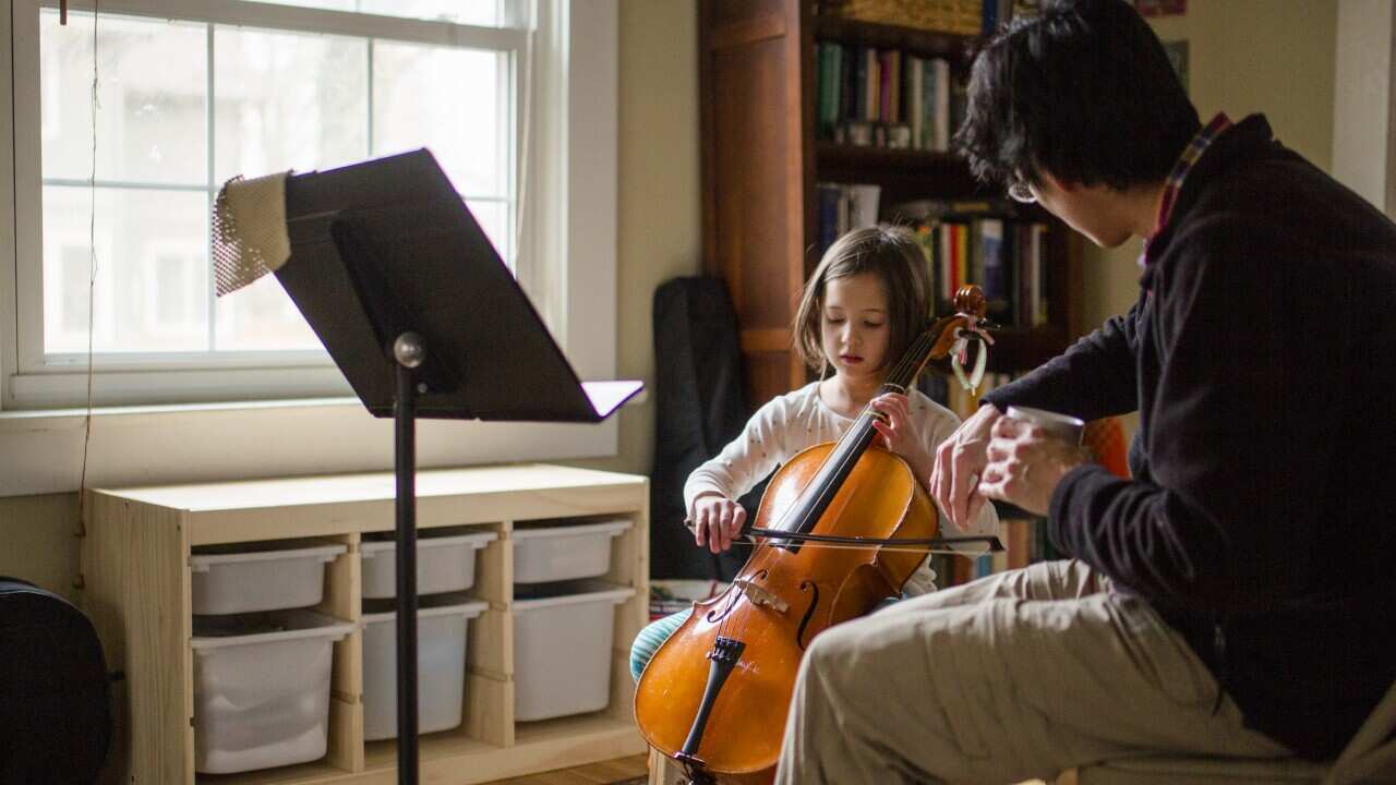 A father helps little girl practice cello at home by window - Australia Explained – Learning an instrument