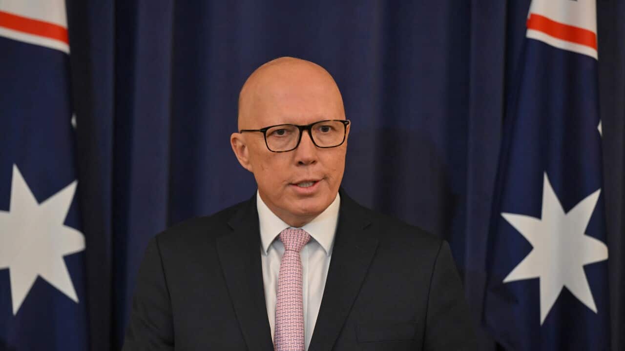 A man in a blue suit next stands in front of two Australian flags.