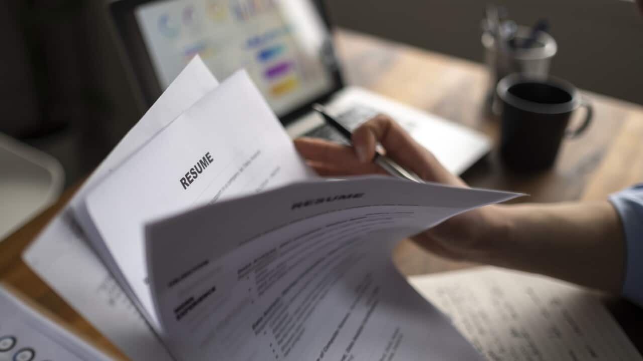 A person's hand flicks through a pile of resumes with a desk and laptop in the background.