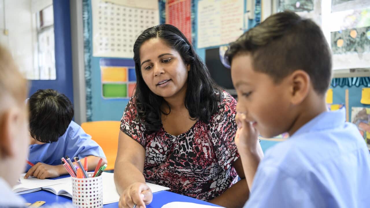 A teacher sits beside a student;s desk and looks over their work in a workbook.