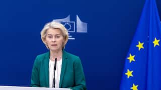 A middle-aged white woman with blonde hair, wearing a teal-coloured blazer jacket and a white shirt. She is standing before a podium with a microphone; the European Union flag is beside her.