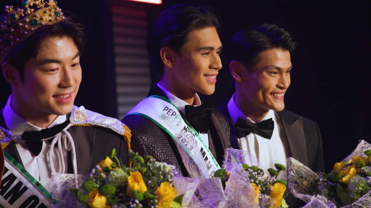 Three young Asian men in tuxedos and pageant ribbon sashes holding bouquets of flowers and smiling. The man on the left wearing a crown and a shiny white cape.