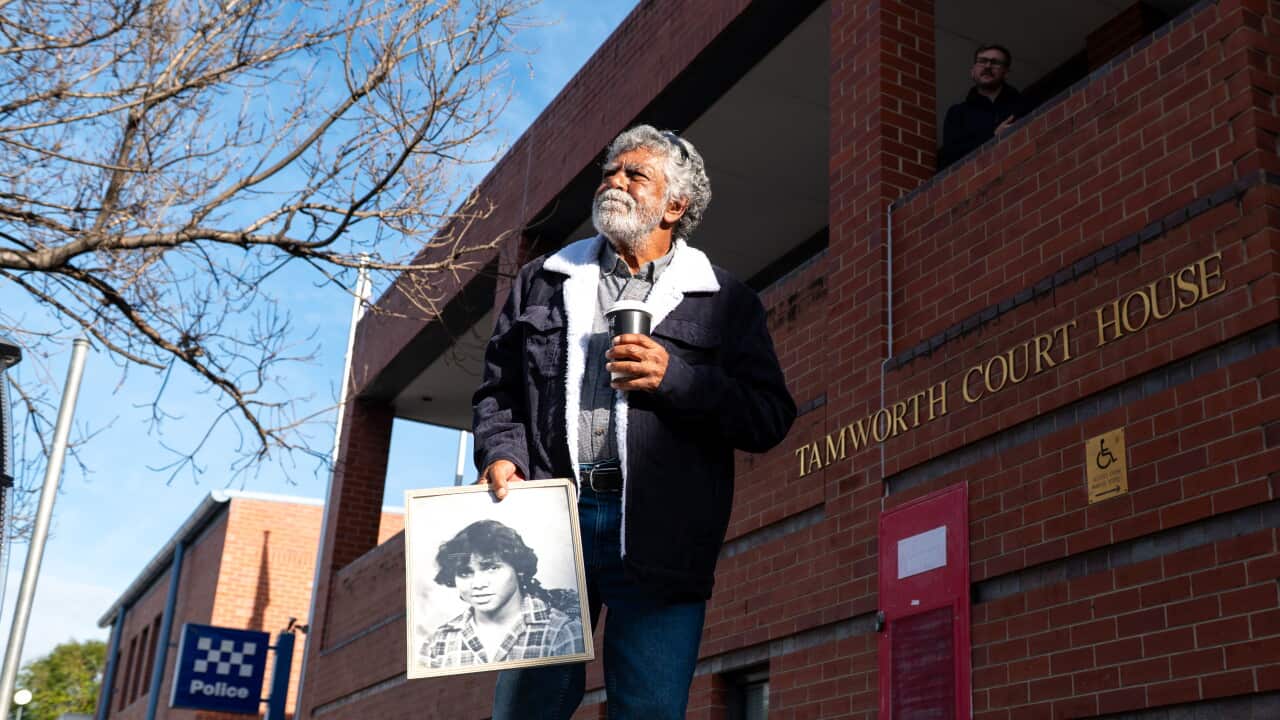 An older First Nations man dressed in a black jacket and blue jeans stands outside a red brick building marked Tamworth Court House. He holds a large, framed black-and-white photograph of a young Aboriginal man in one hand and a coffee cup in the other.