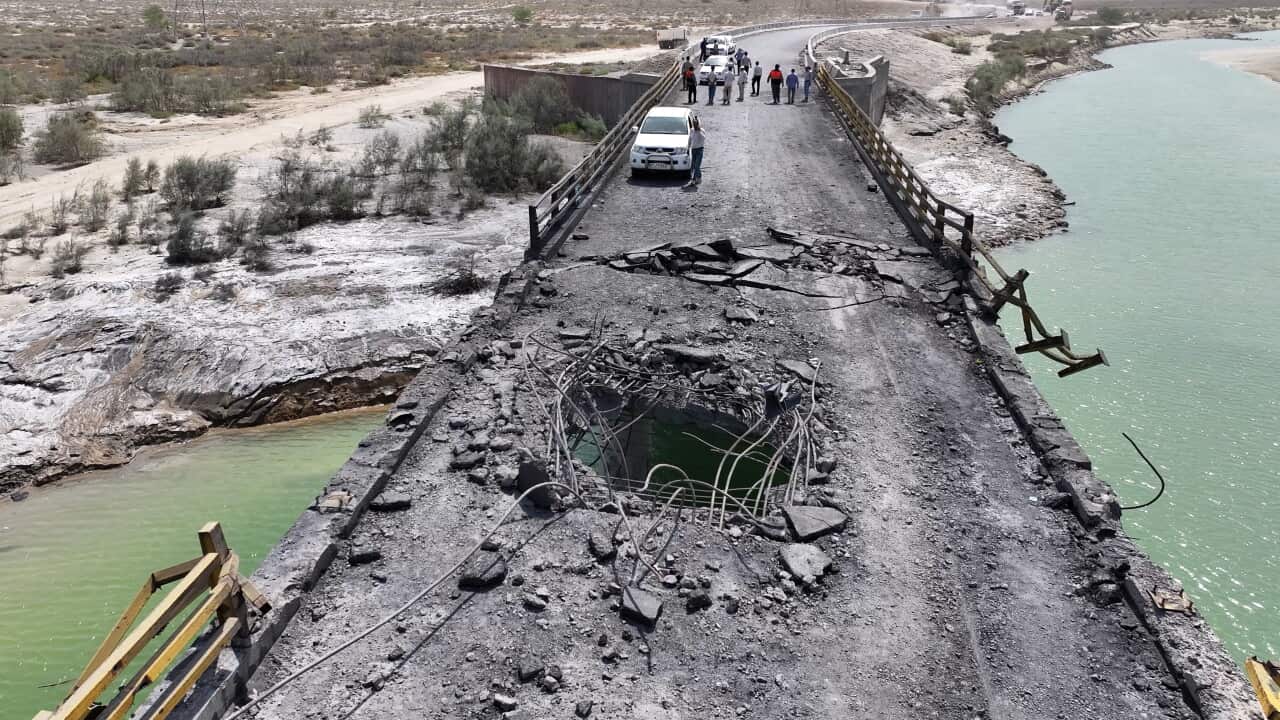 An elevated view shows a heavily damaged asphalt bridge with a large hole exposing rebar in the center roadway, while a white SUV and a group of people stand further down the path near a river bank.