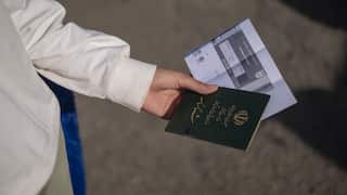 A hand stretches out, holding a green passport with gold writing.