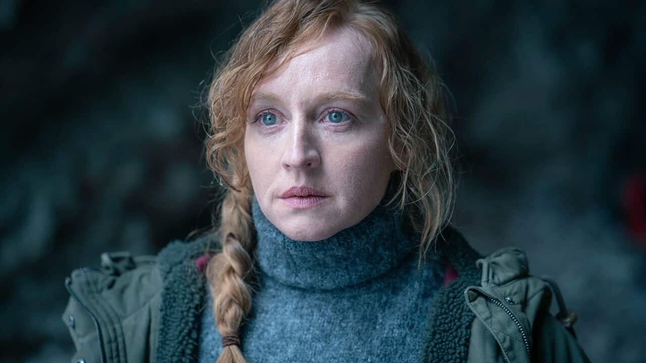 A woman with pale red hair wearing a jumper and coat stars ahead.