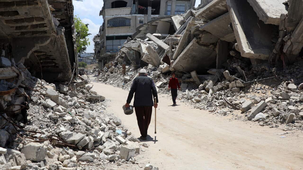 Two people walk down a dirt road lined with destroyed buildings and rubble.