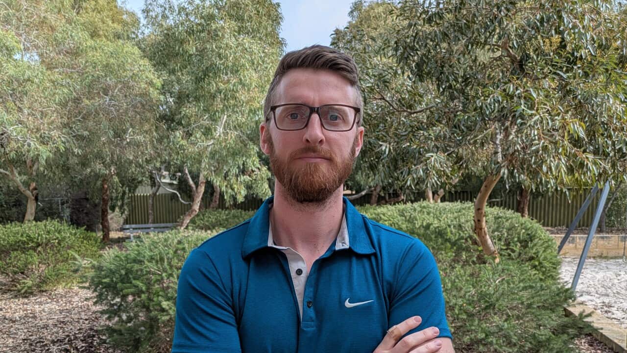 a man in his 30s with ginger hair and a ginger beard in glasses and a blue short sleeved collard shirt stands in a park with his arms crossed