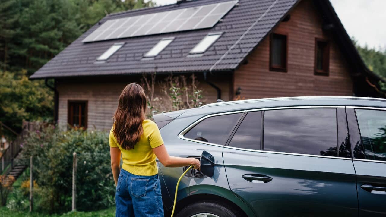 Young woman is charging her electric car, parked in front of a house with solar panels on the roof.