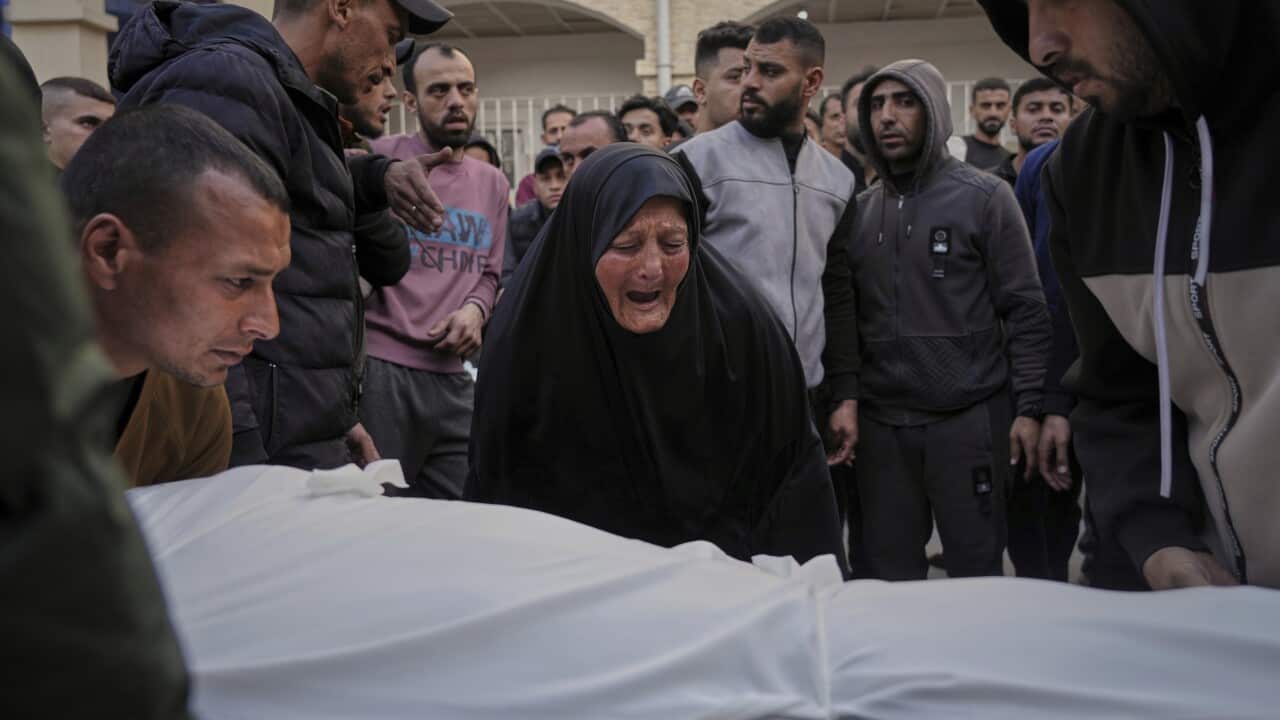A woman mourns over a body covered in a white sheet, with a crowd standing around her.