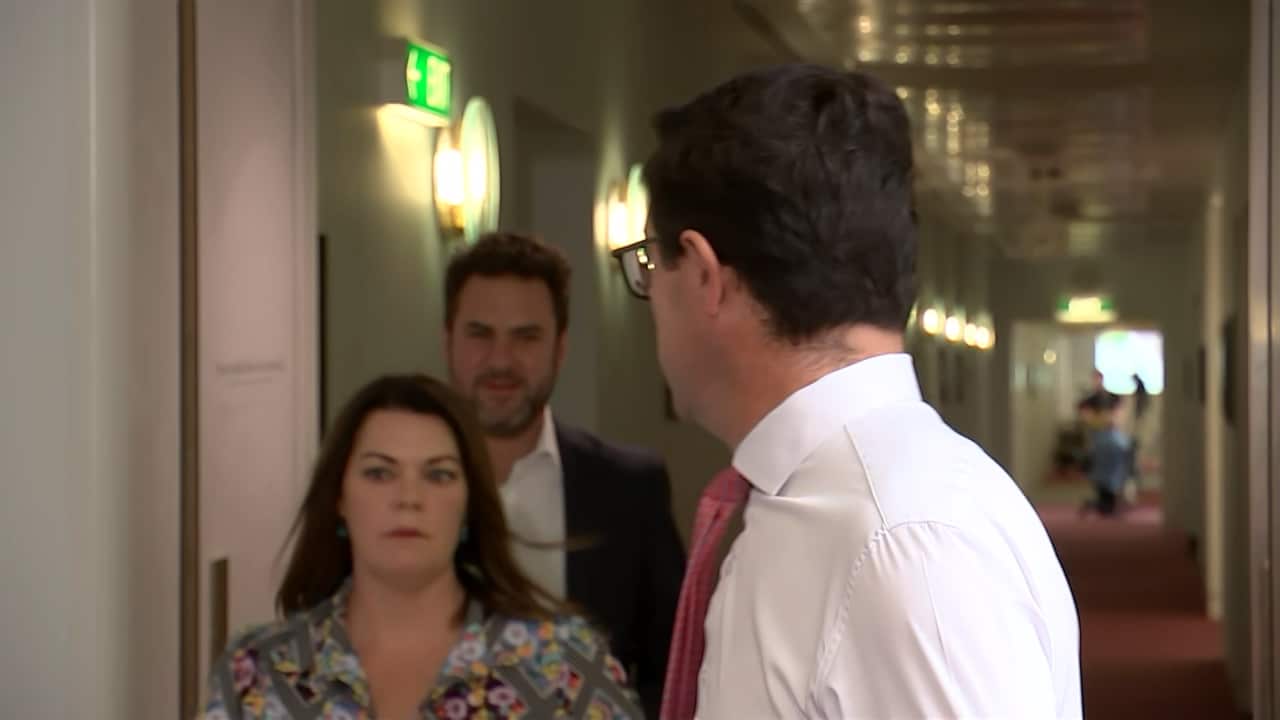 A woman and a man walking past a man who is speaking.