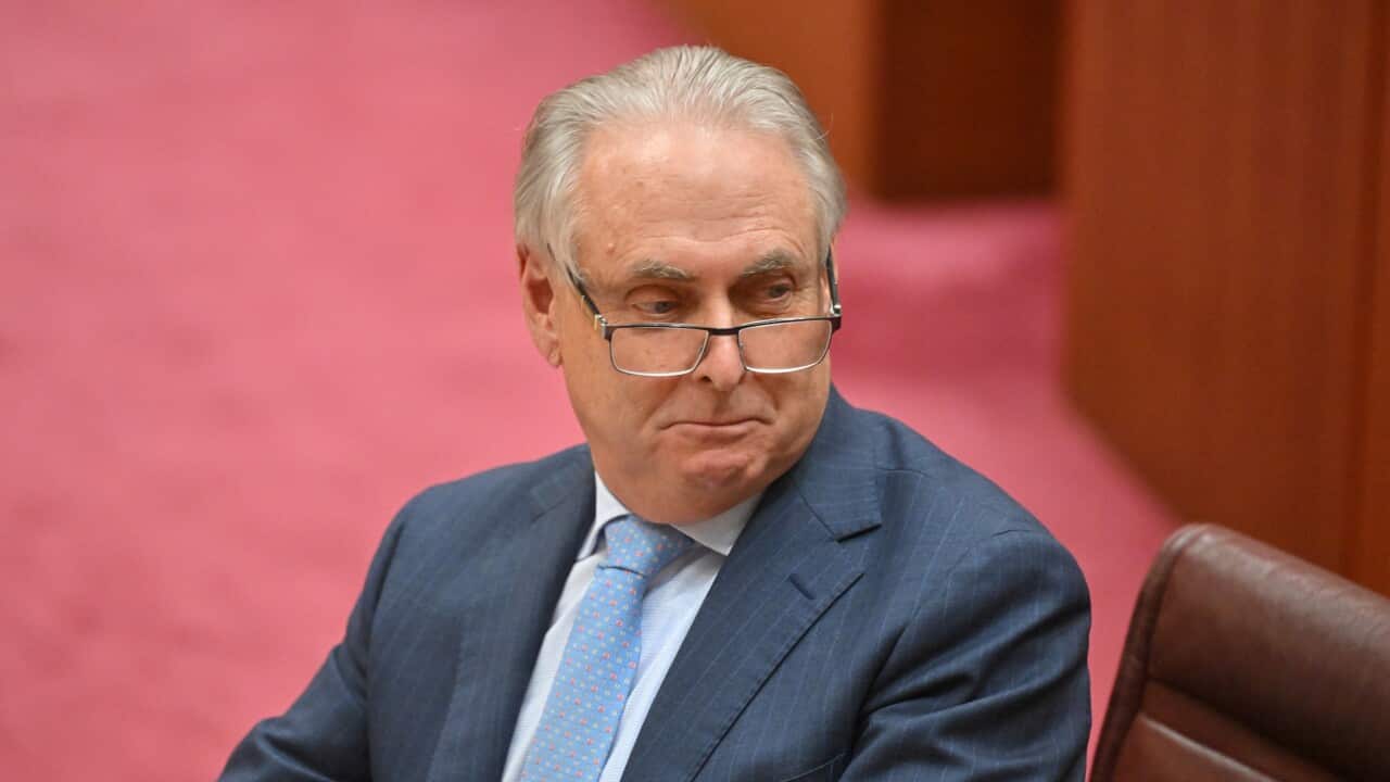 A middle-aged man with grey hair and glasses, wearing a blue suit and light blue patterned tie, looks toward his left with a thoughtful or serious expression.