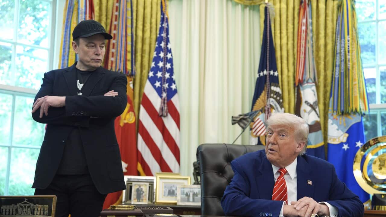 Donald Trump seated in the Oval Office. Elon Musk is standing near him.