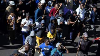 A large crowd of people marches down a street in daylight, many holding long wooden sticks or poles above their shoulders. The group appears to be chanting or shouting as they move together. Most participants are wearing casual clothing, including hoodies, hats, and jackets, while strong sunlight creates deep shadows across the road. The image is photographed from an elevated viewpoint, emphasizing the size and density of the march.