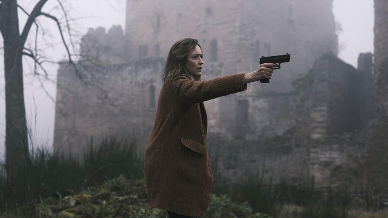 A woman holding a gun, pointed at something or someone unseen, stands in front of a mist-shrouded stone building, with a leafless tree behind her.