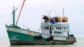 A traditional Thai style fishing boat of the type frequently used by Myanmar Rohingya refugees. It is old and rusty, its hull painted green with a white design on the prow. Colourful clothes are hung over the railings on the upper deck.