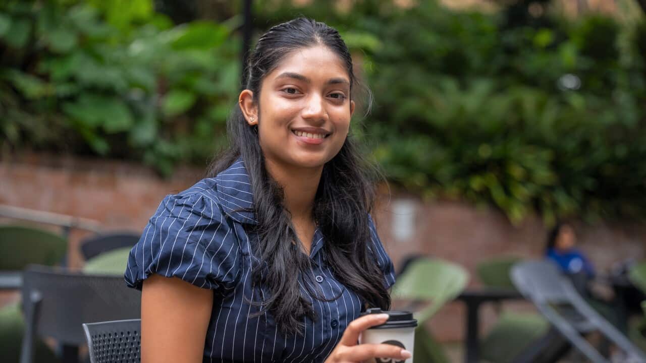 A woman with long black hair smiles as she holds a coffee cup while sitting in a garden.
