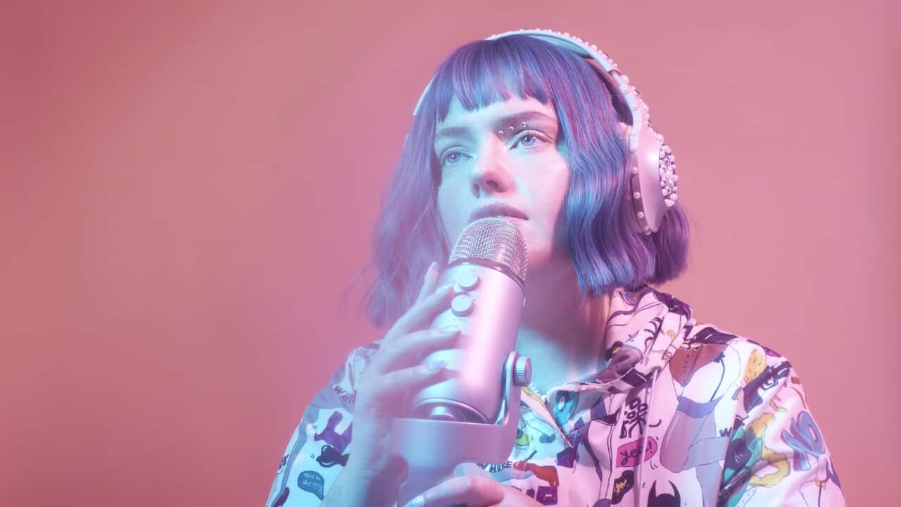 A woman with purple hair wearing pink bedazzled headphones, looking into the distance. She is holding a microphone and bathed in blue light.