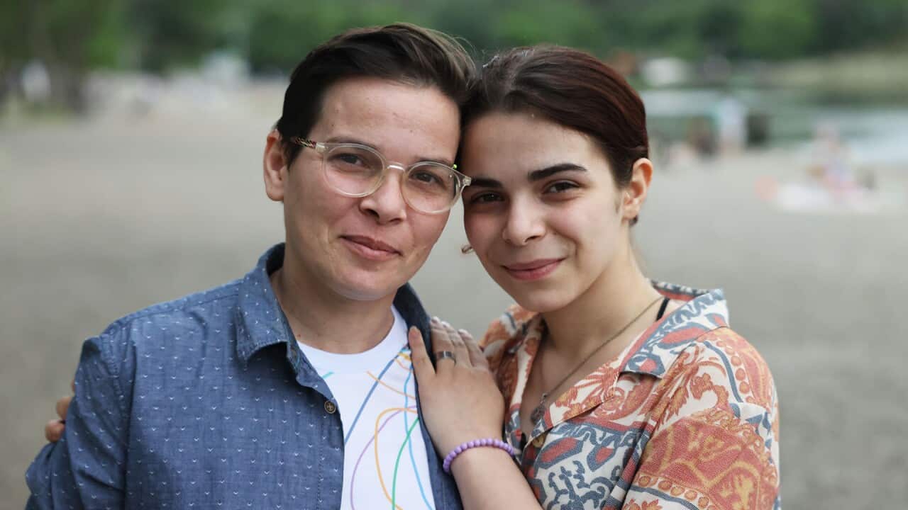 A young woman with dark hair drawn back looking at a camera with her arms around another woman's shoulder. The woman on the left has short hair and eyeglasses.