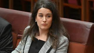 A woman with long dark hair wearing a patterned blazer sits in a formal chamber with red leather seating.