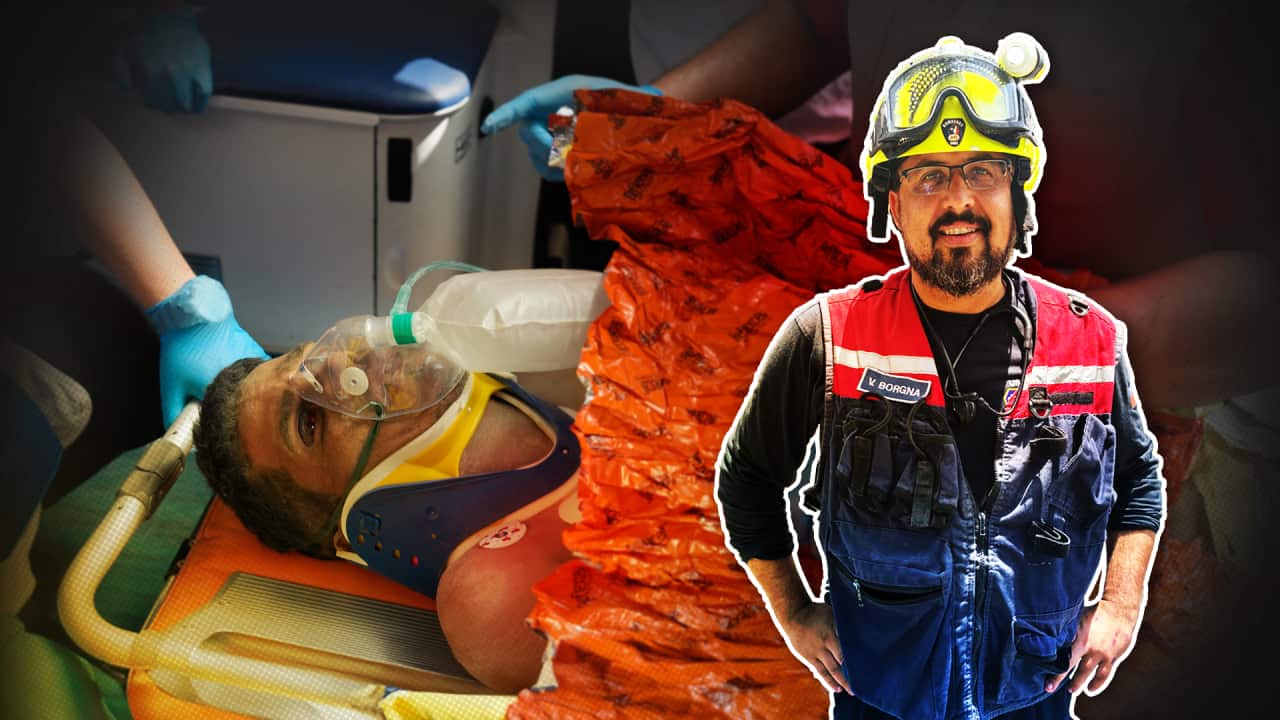A man with a bruised face lies on an orange stretcher, with an oxygen mask to his face. Another man wearing a yellow helmet and rescue uniform stands next to him.