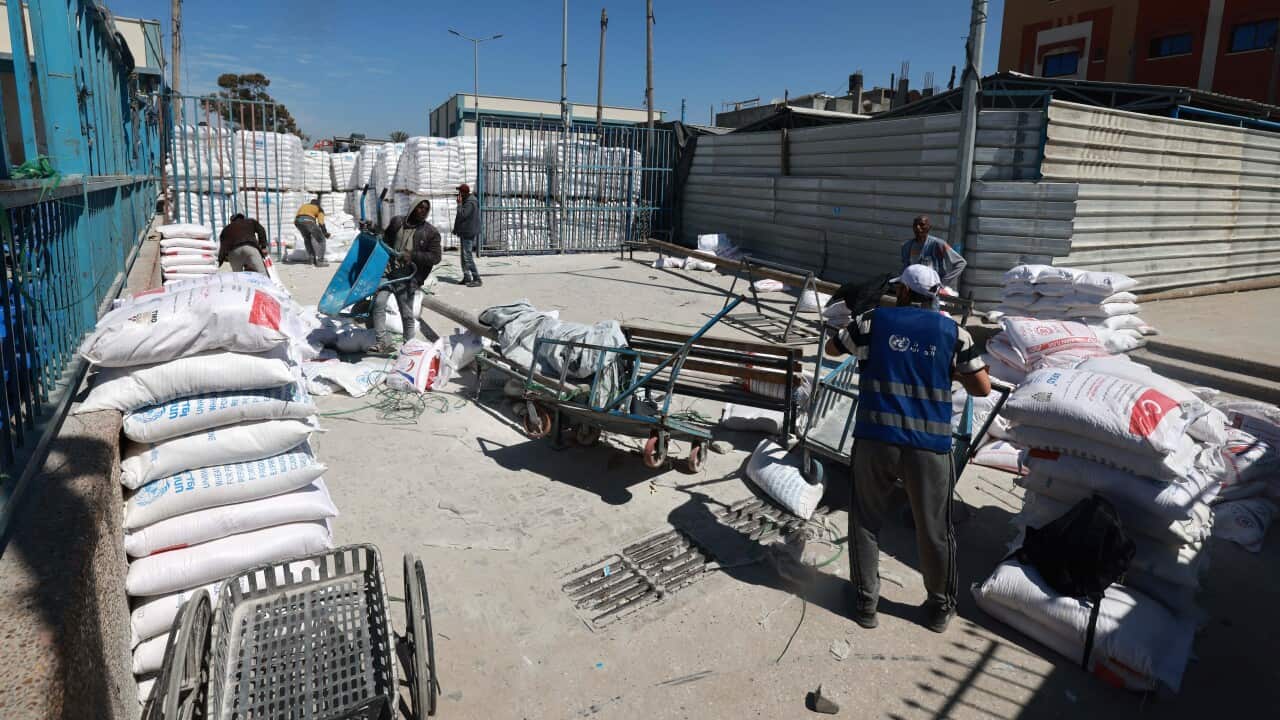 A group of about six people, some in blue vests, appear to be working in a loading dock type area, some of them with wheelbarrows. There are piles of material in large white bags with writing placed loosely around the workers.